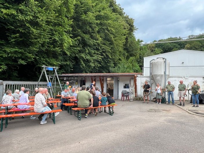 Menschen sitzen an Biertischen und unterhalten sich in einem Hinterhof vor einem Gebäude; Musiker stehen daneben mit Hörnern, während Bäume die Umgebung begrenzen.